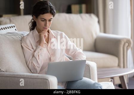 Thoughtful woman spend leisure using laptop sit on couch indoor Stock Photo