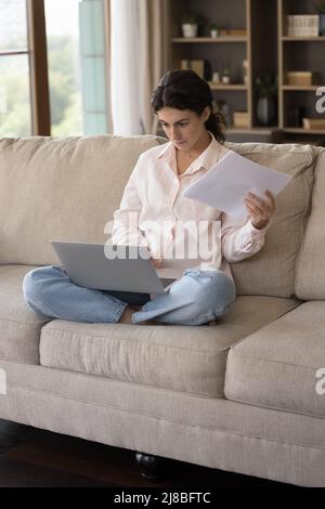 Serious woman sitting on sofa holds documents working on computer Stock Photo