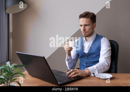 Businessman is working on laptop for new architectural project. Caucasian young man in a shirt and waistcoat sits at a table in the office and works. Considering a development plan Stock Photo