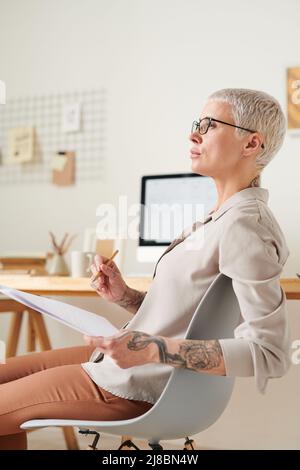 Pensive attractive middle-aged lady with tattoos analyzing papers in own office Stock Photo