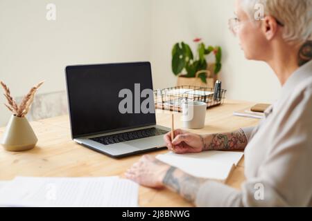 Busy middle-aged woman sitting at desk and making notes in diary while studying to promote business with social media Stock Photo