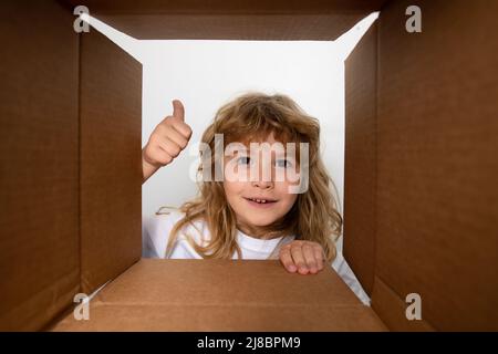 Funny child boy with thumb up is opening gift and looking inside cardboard box. Parcel packaging or unpacking concept. Stock Photo