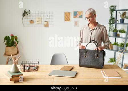 Serious modern middle-aged businesswoman in glasses standing at table and putting folder into bag while going to meeting Stock Photo