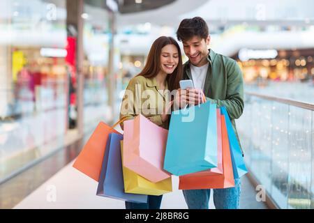 Happy Spouses Doing Shopping And Using Cellphone Standing In Mall Stock Photo