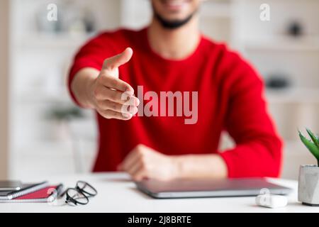 Happy friendly young arabic guy gives hand welcoming at camera at workplace in home office interior, blurred Stock Photo