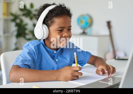 Content inquisitive Black boy in wireless headphones sitting at desk and using laptop while working with tutor Stock Photo
