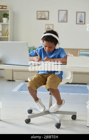 Concentrated black boy in headphones sitting at desk and doing homework with online tutor Stock Photo