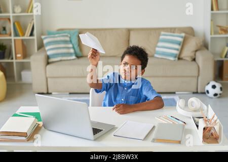 Lazy Black boy with curly hair sitting at desk and playing with paper plane while procrastinating at home Stock Photo