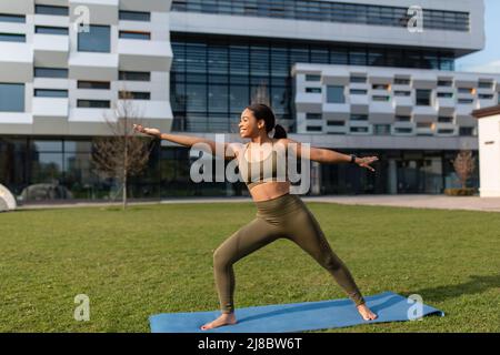 Joyful fit young black woman practicing yoga outdoors, standing in warrior pose, copy space Stock Photo
