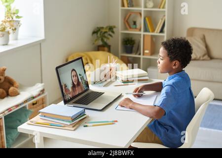 Focused black schoolboy with curly hair sitting at desk and using laptop while listening to tutor during video conference Stock Photo