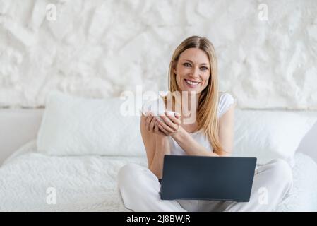 Smiling attractive young blond european woman with cup and laptop sits on bed in white bedroom Stock Photo