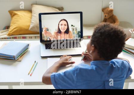 Over shoulder view of black boy sitting at table and looking at laptop screen while connecting with online tutor Stock Photo