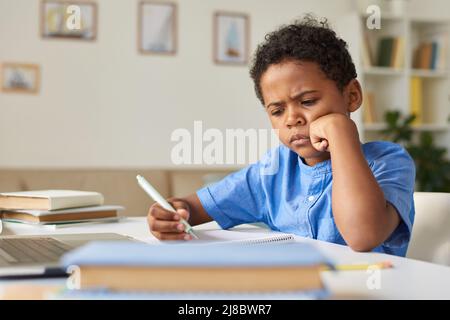 Concentrated African-American schoolboy in blue shirt sitting at desk and learning lessons at home Stock Photo