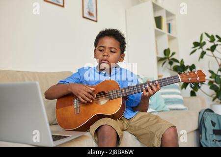 Focused black boy sitting on sofa in living room and watching online video on laptop while learning to play guitar Stock Photo