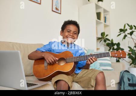 Portrait of cheerful adolescent black boy sitting on sofa in living room and playing guitar according to video lesson Stock Photo