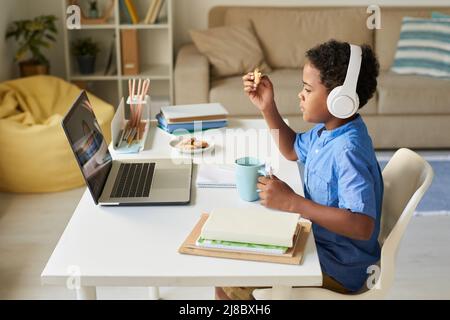 Side view of African-American boy in wireless headphones sitting at home desk and eating cookies while talking to online tutor Stock Photo