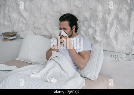 Unhappy millennial european male lies on bed, blows nose at napkin in white bedroom interior Stock Photo