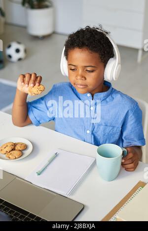 Content cute African-American boy in headphones drinking tea with cookies and watching online video on laptop Stock Photo