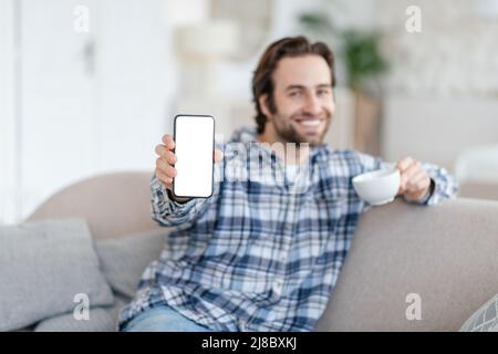 Smiling young caucasian man with stubble sits on sofa with coffee cup, shows phone with blank screen Stock Photo