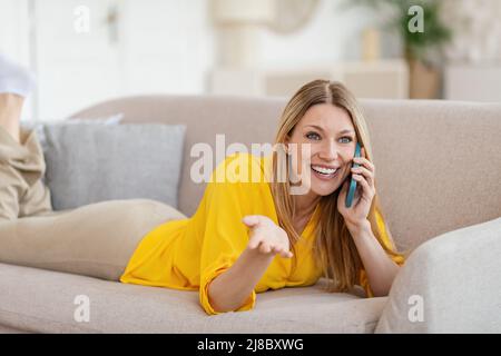 Smiling pretty millennial blond caucasian woman in yellow clothes speaks by phone, lies on sofa Stock Photo