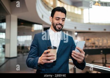 Smiling handsome millennial islamic guy with beard in suit with cup of coffee takeaway looks at phone Stock Photo