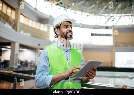 Smiling handsome young muslim male engineer with beard in hard hat and protective uniform with tablet checks project Stock Photo