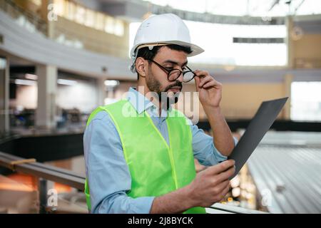 Serious attractive young arab guy engineer with beard in helmet, protective uniform takes off his glasses Stock Photo