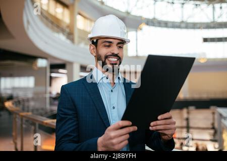 Smiling handsome young arab male engineer, architect or builder with beard in hard hat and suit checks project Stock Photo