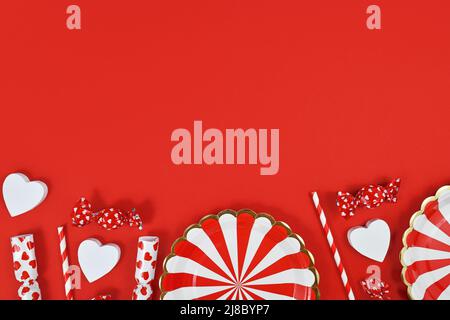 Party flat lay with paper plates, heart ornaments, candy and drinking straws at bottom of red  background with copy space Stock Photo