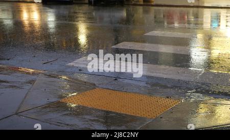 Rain drops on wet asphalt of city street in USA, raindrops falling on zebra crossroad. Cars lights reflection on road in rainy weather. Puddle of water by pavement. Night or twilight dusk atmosphere. Stock Photo