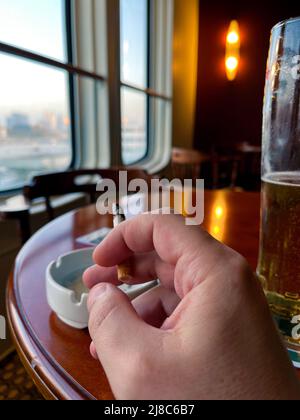 a man's hand holds a cigarette against the background of windows in a beer bar. the concept of the harm of smoking. Stock Photo