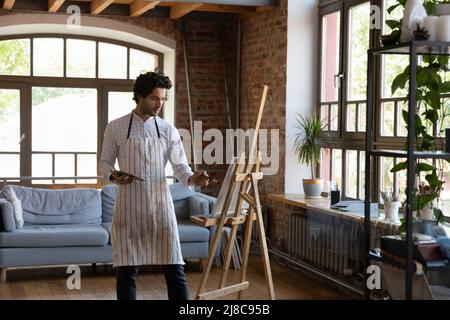 Millennial man painter holding palette painting on canvas in workshop Stock Photo