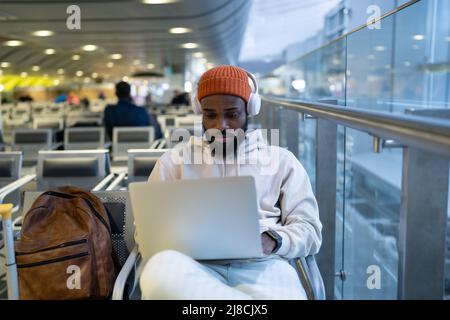 Young hipster black man sitting in airport terminal using laptop wear headphones and listening music Stock Photo
