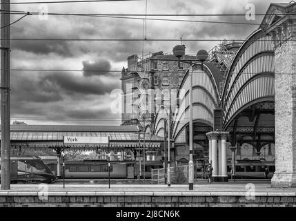 Historic arches are over the entrance to a railway station. Trains stand at platforms and a cloudy sky is above. Stock Photo