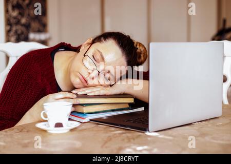 Tired young woman in glasses sleeping on books near laptop at desk in home room. Tiredness from monotonous study and rest at workplace from computer Stock Photo