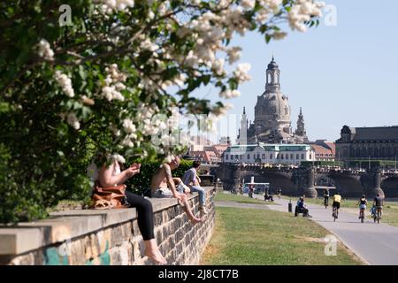 15 May 2022, Saxony, Dresden: Passers-by sit on a wall on the banks of the Elbe River in front of the backdrop of the Frauenkirche church. Photo: Sebastian Kahnert/dpa