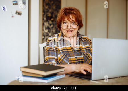 Happy mature lady typing on laptop sitting at desk in room and practicing computer skills. Advanced pensioner using and learning modern technology Stock Photo