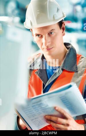 Young inspector or engineer in helmet and construction vest makes journal entries at production facility. Quality control of works. Portrait of Caucasian worker inside room. Stock Photo