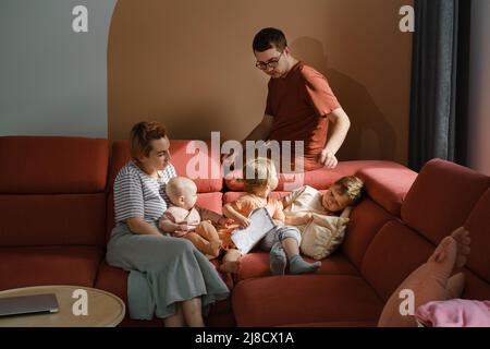 Big family on couch at home. Parents with kids on sofa during sunny day spending free time. Mother and father with three children reading book. Modern Stock Photo