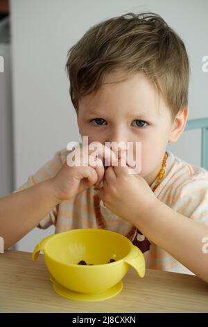 Toddler boy eating happily by the table food by himself in the room. Child having meal. Happy kid having lunch. Little kid with bowl Stock Photo