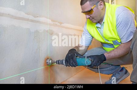 A master electrician in goggles drills a hole in the wall to install an electrical outlet, close up. Stock Photo