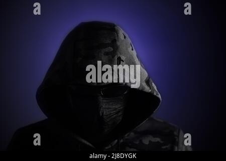 Stylish man in dark hoodie, black glasses and mask dark background in blue light, hacker, attacker and bandit Stock Photo