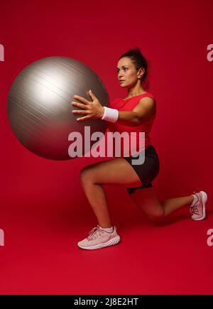Attractive multi-ethnic young athletic woman doing forward lunges while exercising with a fit ball, holding it at chest level, isolated over red backg Stock Photo