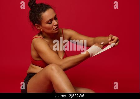 Determined concentrated multiethnic female boxer, sportswoman wrapping her arm with elastic bandages for boxing, isolated on red background with adver Stock Photo