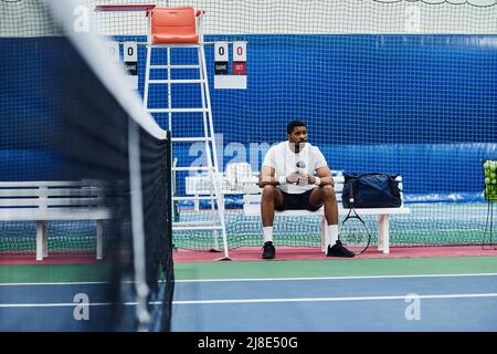 Wide angle shot of professional black sportsman sitting on bench in tennis court and holding water bottle, copy space Stock Photo