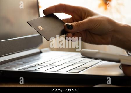 A girl holds a credit bank card in her hands near a black laptop in an office near a window, online payment, internet payment Stock Photo