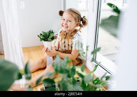 Smiling with teeth little girl sitting on a window sill. She's holding a spray bottle, looking at the camera. She is taking care of plant seedlings Stock Photo