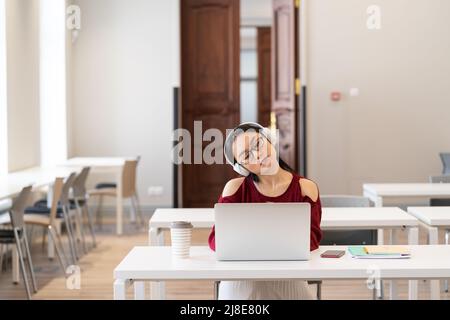 Tired Asian student girl in headphones doing gymnastics while studying online in modern classroom Stock Photo