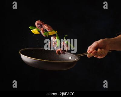 A frying pan with seafood, lemon slices and vegetables in the hand of a professional chef on a black background. Levitation. Advertising, banner. Ther Stock Photo