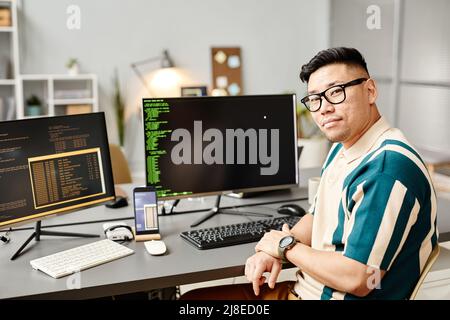 Portrait of Asian IT developer looking at camera against programming code on computer screen in office interior, copy space Stock Photo